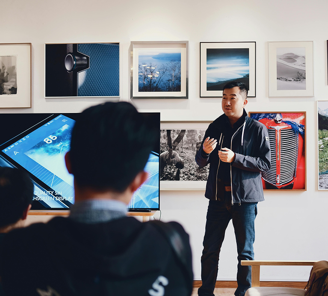 Asian man in casual clothing speaking at a gallery event next to large tv screen showing presentation